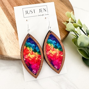 May include: Rainbow tie-dye wooden earrings with silver hooks. The earrings are shaped like a teardrop and have a wood grain finish. The earrings are handmade and are a unique and colorful accessory.