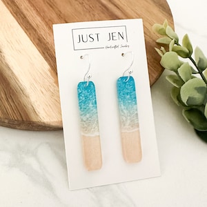 May include: A pair of dangle earrings with a beach scene design. The earrings are made of resin and feature a blue ocean, white waves, and a light brown sandy beach. The earrings are hanging from silver hooks.