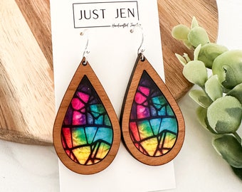 May include: Rainbow stained glass design wood earrings with silver hooks. The earrings are teardrop shaped and have a wood frame. The text "JUST JEN - Handcrafted Jewelry" is on the white card behind the earrings.