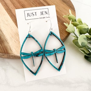 May include: Teal blue dragonfly earrings with a wood grain finish. The earrings are shaped like a teardrop and have a silver hook.