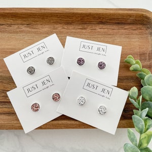 May include: Four pairs of stud earrings on white cards, each with the text "JUST JEN". The earrings have a round, textured design in shades of gray, purple, rose gold, and white, set in silver-colored metal. The earrings are displayed on a wooden tray.