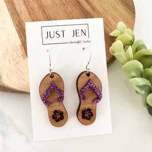 May include: A pair of wooden earrings shaped like flip flops with purple glitter accents and a hibiscus flower design. The earrings are on a white card with the text "JUST JENI - Handcrafted Jewelry".