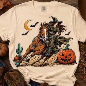 May include: A cream-colored short-sleeve t-shirt featuring a graphic of a witch in a pointed hat riding a brown horse. The scene includes a jack-o'-lantern, a crescent moon, bats, and a cactus. The shirt has a Comfort Colors brand label.