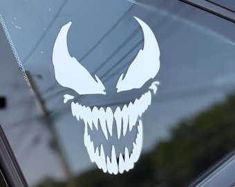 May include: White vinyl decal of a monster face with sharp teeth and pointed eyes, applied to a car window. The design features a menacing grin and is likely a decorative car accessory.
