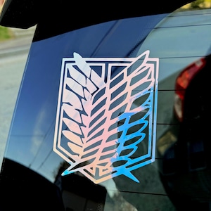 May include: A holographic decal of the Attack on Titan Survey Corps emblem, featuring a stylized wing design within a shield shape. The decal is affixed to a car window, reflecting a spectrum of colors.