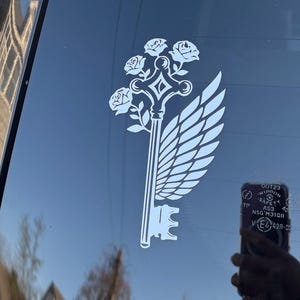 May include: White decal featuring a key design with roses, a wing, and a decorative handle. The key is a symbol of unlocking secrets or possibilities. The decal is applied to a dark surface, likely a car window.