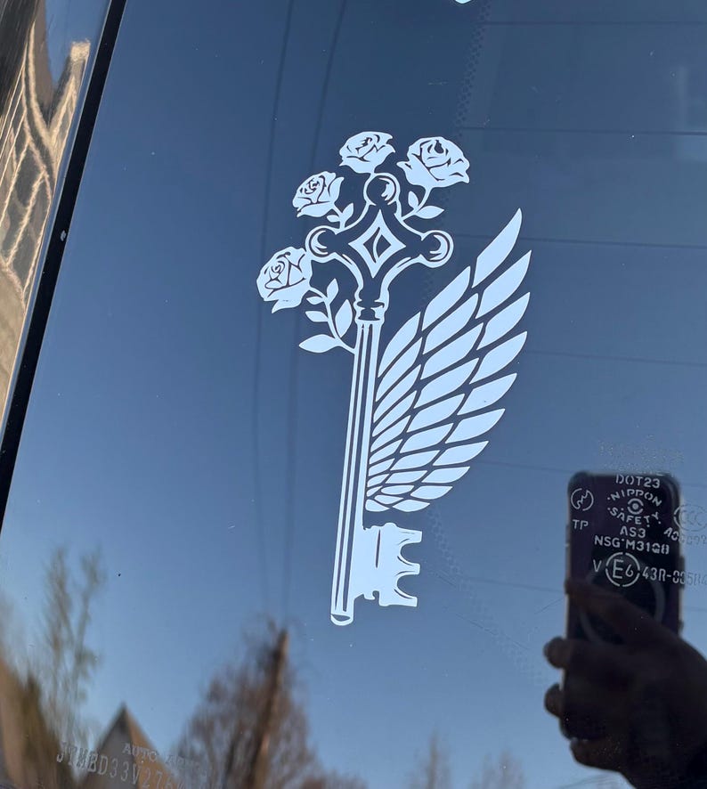 May include: White decal featuring a key design with roses, a wing, and a decorative handle. The key is a symbol of unlocking secrets or possibilities. The decal is applied to a dark surface, likely a car window.