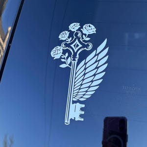 May include: A white decal of a key with a wing and roses on a car window. The key design features a key head, a stem, and a decorative top with roses. A wing is on the right side of the key.