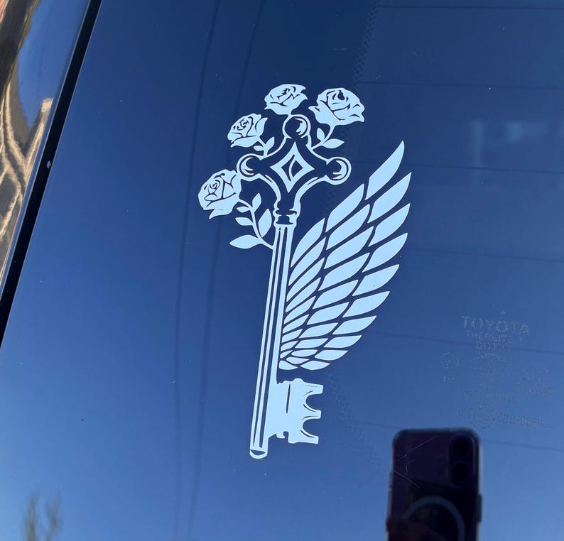 May include: A white decal of a key with a wing and roses on a car window. The key design features a key head, a stem, and a decorative top with roses. A wing is on the right side of the key.