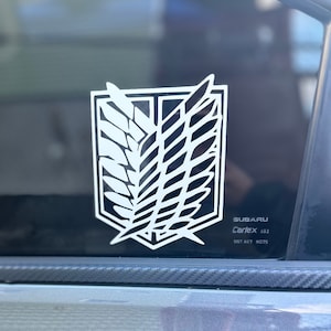 May include: A white vinyl sticker of the Attack on Titan Wings of Freedom emblem on a black car window. The sticker is on a Subaru Carlex 452 with the DOT 467 H075 sticker below it.