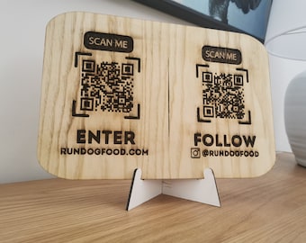Op de afbeelding: Houten bord met twee QR-codes, de tekst "SCAN ME", "ENTER RUNDOGFOOD.COM" en "FOLLOW @RUNDOGFOOD". Het bord wordt ondersteund door een witte standaard. Het hout heeft een lichtbruine nerf.