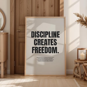 May include: A framed print with the text "DISCIPLINE CREATES FREEDOM" in bold black letters. The print is in a light-coloured wooden frame and is displayed against a neutral-toned wall. The room has a natural, minimalist aesthetic.