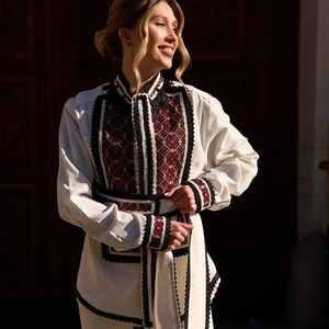 May include: A white blouse and pants set with intricate embroidery. The blouse features a high collar and cuffs with black and red geometric patterns. The outfit includes a matching sash and is a traditional style.