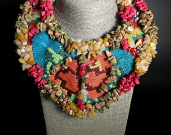 May include: A colorful beaded necklace displayed on a gray bust. The necklace features a central design with blue, orange, and green patterns, surrounded by a border of small, colorful stones and beads in shades of red, yellow, and green.