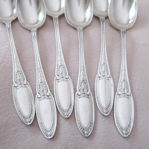 May include: Seven silver spoons with ornate handle detailing. The spoons have a classic design with a polished finish. The spoons are arranged on a light pink surface, showcasing their reflective quality. Ideal for serving or display.