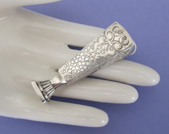May include: A silver seal with an ornate design. The seal is in the shape of a rectangle with a rounded top. The design is intricate and includes floral motifs.
