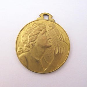 May include: A circular gold-toned metal pendant featuring a relief sculpture of a person with wavy hair playing a stringed instrument. The pendant has a small loop at the top for attachment, with a smooth, polished finish and detailed facial features.