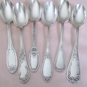 Puede incluir: Un conjunto de siete cucharas de plata con mangos ornamentados, dispuestas sobre un fondo rosa. Cada cuchara presenta un diseño diferente y detallado, reflejando la luz. El conjunto destaca el brillo y el estilo vintage.