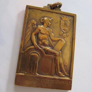 K&ouml;nnte beinhalten: Eine Bronzemedaille mit einer gefl&uuml;gelten Figur, die eine Schriftrolle h&auml;lt. Die Medaille ist mit "EXPOSITION INTERNATIONALE DE LEIPZIG" beschriftet.