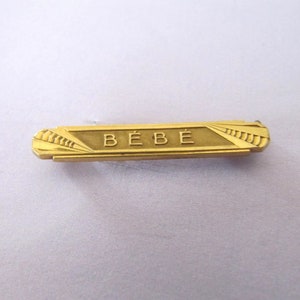 May include: A gold-colored metal pin with the word "BÉBÉ" engraved on it.