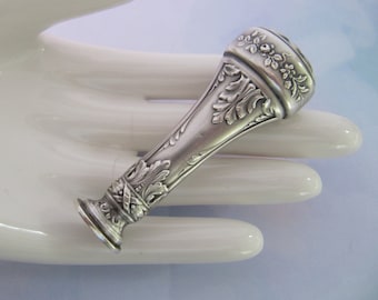 May include: A silver seal with an ornate design, featuring floral and leaf motifs. The seal is in the shape of a cylinder with a rounded top and a flat base.