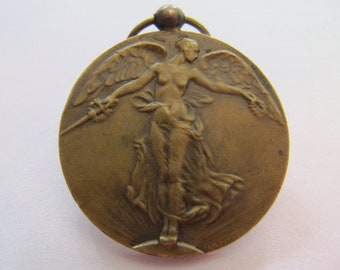 Könnte beinhalten: Eine Bronzemedaille mit einem Reliefbild einer geflügelten Figur, die ein Schwert und einen Kranz hält. Die Figur steht auf einem Sockel und die Medaille hat oben eine Schlaufe zum Aufhängen.