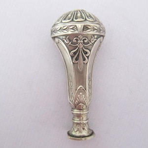 May include: A silver-colored antique wax seal with ornate floral and leaf designs. The seal has a rounded top with a detailed pattern, a flared body, and a decorative base. The overall design is symmetrical and elegant.