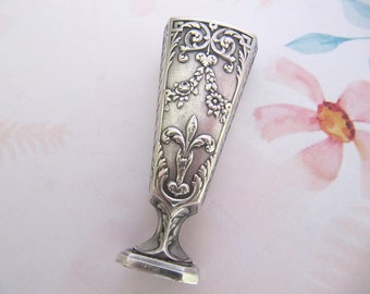 May include: Silver seal with an ornate design featuring a fleur-de-lis and floral motifs. The seal has a square base and a rectangular top.