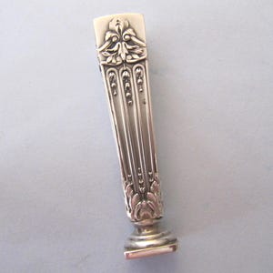 Könnte beinhalten: Ein silberfarbenes antikes Wachssiegel mit aufwendigen Blumen- und Blattdetails. Die rechteckige Basis ist zum Bedrucken bestimmt. Der Griff weist vertikale Linien und dekorative Elemente auf, was auf einen Vintage-Artikel hindeutet.