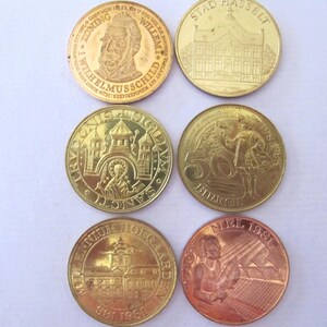 May include: A collection of six gold-colored commemorative coins. The coins feature various designs, including portraits, buildings, and historical figures. Some coins have text inscriptions, such as "STAD HASSELT" and "NIEL 1981".