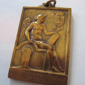 K&ouml;nnte beinhalten: Eine Bronzemedaille mit einer gefl&uuml;gelten Figur, die auf einem Thron sitzt und eine Schriftrolle h&auml;lt. Die Medaille tr&auml;gt den Text "Exposition Internationale de Li&egrave;ge" am unteren Rand.
