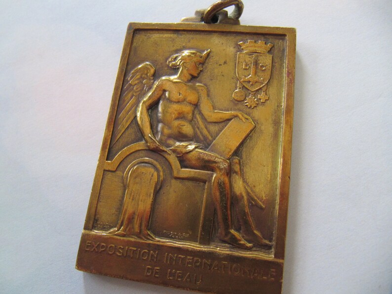 K&ouml;nnte beinhalten: Eine Bronzemedaille mit einer gefl&uuml;gelten Figur, die auf einem Thron sitzt und eine Schriftrolle h&auml;lt. Die Medaille tr&auml;gt den Text "Exposition Internationale de Li&egrave;ge" am unteren Rand.