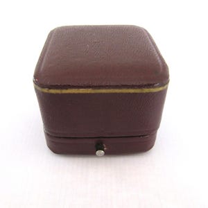 Puede incluir: Una pequeña caja de joyas vintage de color marrón oscuro. La caja tiene forma rectangular con bordes redondeados y un ribete dorado. Se puede ver un pequeño cierre plateado en la parte delantera.