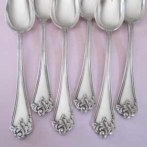 Può includere: Un set di sette cucchiai d'argento disposti su una superficie lilla chiaro. Ogni cucchiaio ha una coppa arrotondata e un manico lungo e sottile con un motivo floreale decorativo alla base. I cucchiai sono in metallo lucido e riflettente.