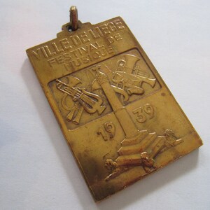 K&ouml;nnte beinhalten: Eine goldfarbene Metallmedaille mit dem Text "VILLE DE LIEGE FESTIVAL DE MUSIQUE 1939" und einem Design von Musikinstrumenten.