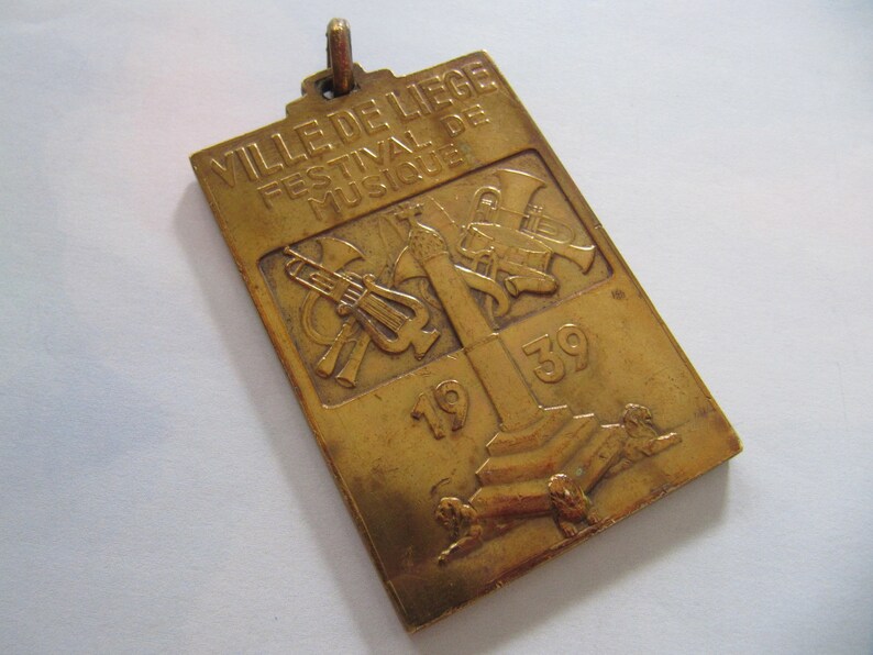 K&ouml;nnte beinhalten: Eine goldfarbene Metallmedaille mit dem Text "VILLE DE LIEGE FESTIVAL DE MUSIQUE 1939" und einem Design von Musikinstrumenten.