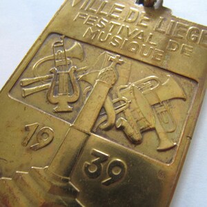 K&ouml;nnte beinhalten: Ein goldfarbenes Metallabzeichen mit dem Text "VILLE DE LIEGE FESTIVAL DE MUSIQUE" und den Zahlen "1939". Das Abzeichen zeigt ein Design aus Musikinstrumenten, darunter eine Trompete, eine Tuba und eine Leier.