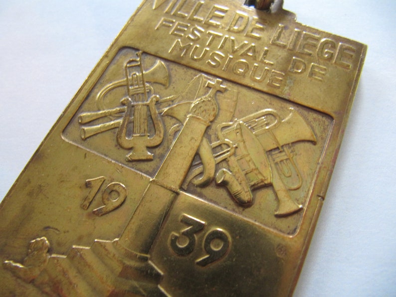 K&ouml;nnte beinhalten: Ein goldfarbenes Metallabzeichen mit dem Text "VILLE DE LIEGE FESTIVAL DE MUSIQUE" und den Zahlen "1939". Das Abzeichen zeigt ein Design aus Musikinstrumenten, darunter eine Trompete, eine Tuba und eine Leier.