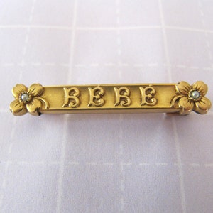 Puede incluir: Broche de barra dorada con la palabra "BEBE" en letras ornamentadas, acentuada con dos diseños de flores con falsas perlas en el centro.