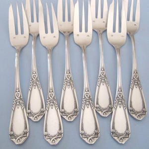 May include: A collection of eight silver forks with three tines each. The handles feature an ornate design with heart and scroll details. The forks are arranged on a light blue surface, showcasing their elegant, vintage style.