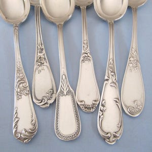 Può includere: Sei cucchiai d'argento antichi con disegni floreali e a volute ornati. I cucchiai hanno una superficie lucida e riflettente e diversi motivi sui manici. I cucchiai sono disposti su uno sfondo azzurro, mettendo in mostra i loro dettagli intricati e il fascino vintage.