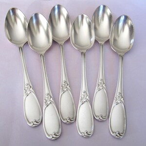 May include: Six silver spoons with ornate handles. The spoons are arranged in a row, with the handles pointing towards the viewer.