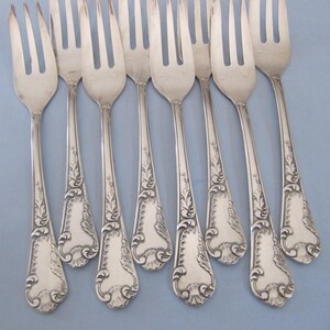 Puede incluir: Un conjunto de ocho tenedores plateados con tres púas cada uno. Los tenedores tienen mangos decorativos con un diseño floral. Los tenedores están colocados sobre una superficie azul claro.