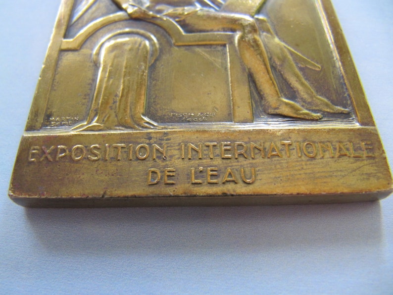 K&ouml;nnte beinhalten: Eine Bronzemedaille mit einem Relief einer sitzenden Figur. Die Medaille ist mit "Exposition Internationale de L'Eau" beschriftet.