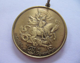 Könnte beinhalten: Eine Bronzemedaille mit der Darstellung des Heiligen Georg, der einen Drachen tötet. Die Medaille ist mit dem Text "St. Landelijke 19 99 Rijvereniging" beschriftet und trägt ein kleines Logo.