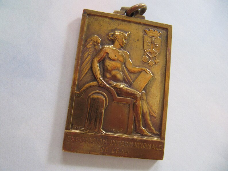 K&ouml;nnte beinhalten: Eine Bronzemedaille mit einer gefl&uuml;gelten Figur, die eine Schriftrolle h&auml;lt. Die Medaille ist mit "EXPOSITION INTERNATIONALE DE LEIPZIG" beschriftet.