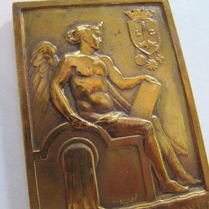 May include: A bronze plaque featuring a relief sculpture of a winged figure seated on a throne, holding a scroll. The figure is wearing a crown and has a muscular physique. The plaque also features a coat of arms with the letters "L.C." and a star.