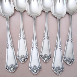 Peut inclure: Six cuillères en argent avec des manches ornés et détaillés et des cuillères ovales sont disposées en rangée. Les cuillères ont une surface polie et réfléchissante, et les manches présentent un motif floral décoratif. Les cuillères sont posées sur un fond rose doux.