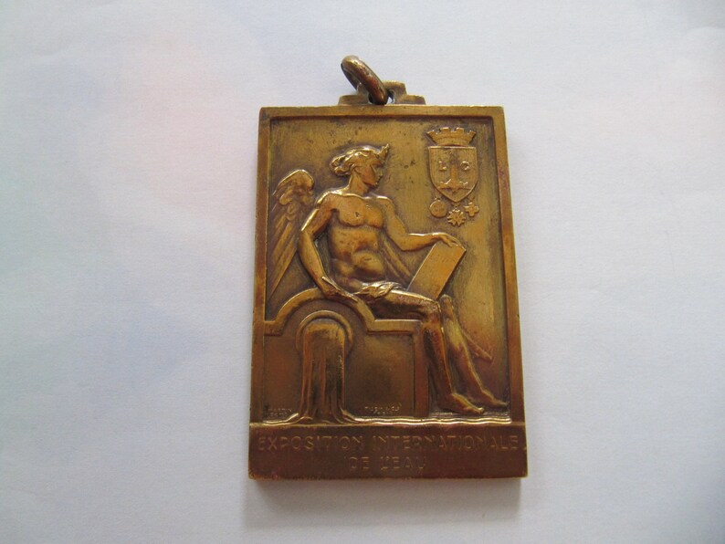 K&ouml;nnte beinhalten: Eine goldfarbene Metallmedaille mit einer Reliefskulptur einer gefl&uuml;gelten Figur, die eine Schriftrolle h&auml;lt. Die Medaille ist mit "EXPOSITION INTERNATIONALE DE LYON" beschriftet.