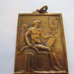 K&ouml;nnte beinhalten: Eine goldfarbene Metallmedaille mit einer gefl&uuml;gelten Figur, die eine Schriftrolle h&auml;lt. Die Medaille zeigt ein Wappen mit den Buchstaben "L.C." und einer Krone.